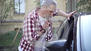 Adult Caucasian grey-haired man approaching to car outdoors stops having chest pain. Portrait of ill guy having heart