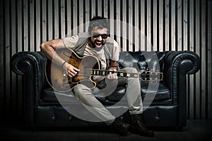 Adult caucasian guitarist portrait playing electric guitar sitting on vintage sofa. Music singer concept on couch and