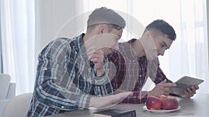 Adult Caucasian man giving tablet to his twin brother, sibling giving him book. Two identical twins resting on weekends