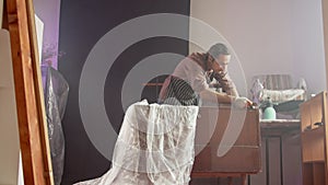 An adult Caucasian man in a special protective apron is restoring an old chest of drawers in a working workshop. Small