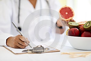 Adult caucasian nutritionist doctor in white coat at table makes notes in clipboard, shows grapefruit