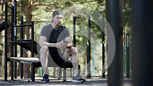 Adult caucasian strong sportsman is taking a rest on a bench and drinks water out of the shaker at the gym at the park