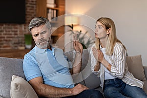 Adult caucasian wife asks and prays with gesture at sad husband with beard, man ignores lady