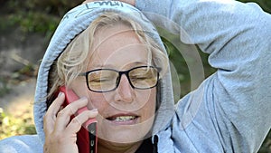 Adult caucasian woman with glasses having fun talking on smartphone with red cover. Blond middle aged female is resting.