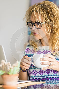 Adult caucasian woman look laptop computer at home drinking with a cup - pretty blonde female people use technology for work or