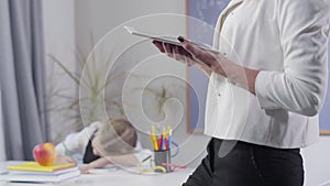 Adult caucasian woman standing with tablet as little pretty schoolgirl writing in exercise book. Blurred child studying