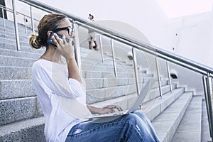 Adult caucasian woman working with technology and modern devices like phone and laptop outdoor sitting on a stairs in urban city