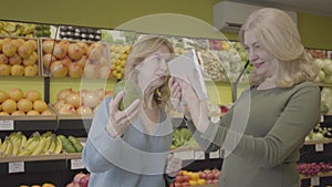 Adult Caucasian women checking fruit origin using tablet. Positive housewives buying pomelo in grocery. Lifestyle