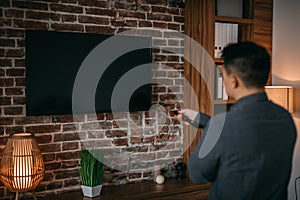 Adult chinese man with remote control turns on tv with blank screen in home room interior, back