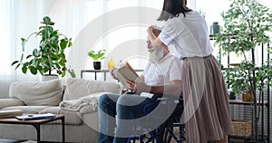 Adult daughter combing hair of disabled old father reading book on wheelchair