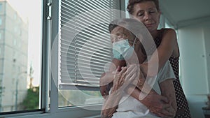 Adult daughter embracing with loving senior mother 90 years in mask standing at window, relatives from different