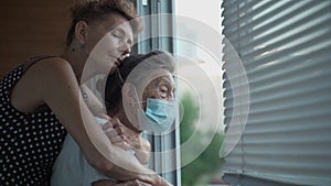 Adult daughter embracing with loving senior mother 90 years in mask standing at window, relatives from different