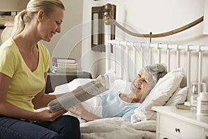 Adult Daughter Reading Newspaper To Senior Female Parent In Bed At Home