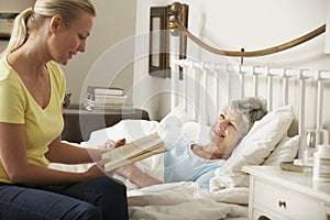 Adult Daughter Reading To Senior Female Parent In Bed At Home