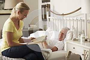 Adult Daughter Reading To Senior Male Parent In Bed At Home