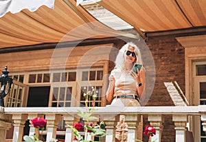 Adult elegant blonde woman in sunglasses and beige clothes using mobile phone at house backyard, in summer city street cafe