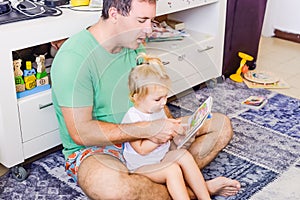 Adult father with toddler daughtersitting at home and reading book story. Little girl sitting on father lap. Family, children educ