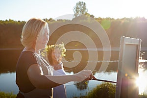 Adult female artist painting picture near water of river or lake in nature and girl in white sundress and flower wreath