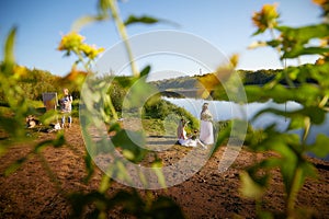 Adult female artist painting picture near water of river or lake in nature and girls in white sundress and flower wreath