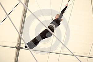 An adult female hangs on a flying trapeze at an indoor gym. The woman is an amateur trapeze artist