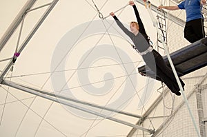 An adult female hangs on a flying trapeze at an indoor gym. The woman is an amateur trapeze artist