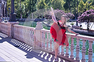 Adult female Hispanic classical ballet dancer in red tutu doing figures next to a stone railing in the middle of a plaza on a