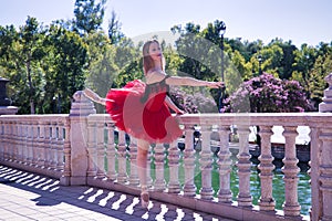 Adult female Hispanic classical ballet dancer in red tutu doing figures next to a stone railing in the middle of a plaza on a