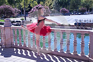 Adult female Hispanic classical ballet dancer in red tutu doing figures next to a stone railing in the middle of a plaza on a