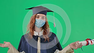adult female student in medical mask and an academic gown and hat with diploma spreads arms to side on green background