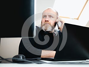 Adult focused man calling by smartphone during remote work on laptop and using two monitors from home