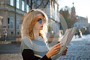 Adult girl in blue sunglassed is looking at the paper map and searching for direction early in the morning in ancient