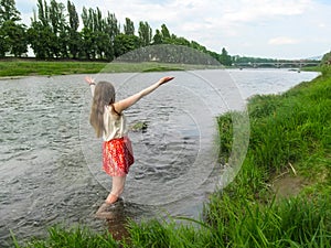 Adult girl with long blond hair splashes in the water of Uzh River - back view. A young woman enjoys spring freshness of water,