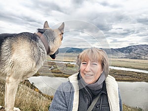 Adult girl with shepherd dog taking selfie near water of lake or river in mountain. Middle aged woman and big pet on