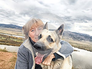 Adult girl with shepherd dog taking selfie near water of lake or river in mountain. Middle aged woman and big pet on