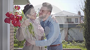 Adult grandson visiting his grandmother, bringing her bouquet of tulips. The bearded man hugging his granny. People