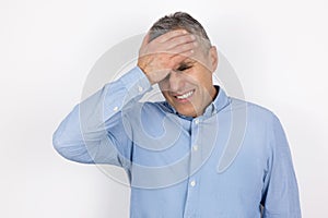 Adult handsome man with grey hair wearing blue shirt touching forehead with hand feeling sorry standing on isolated white