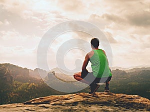 Adult hiker in black shorts and green singlet sit on mountain edge. Man enjoying view