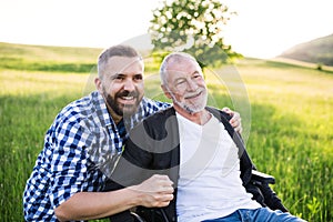 An adult hipster son with senior father in wheelchair on a walk in nature at sunset, laughing.