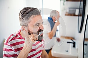 An adult hipster son and senior father brushing teeth indoors at home.