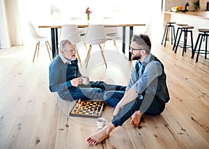 An adult hipster son and senior father sitting on floor indoors at home, playing chess.