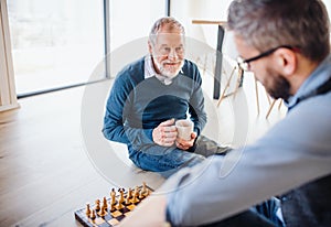 An adult hipster son and senior father sitting on floor indoors at home, playing chess.