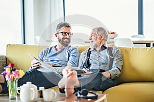 An adult hipster son and senior father sitting on sofa indoors at home, talking.