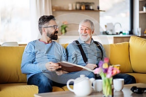 An adult hipster son and senior father sitting on sofa indoors at home, talking.