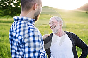 An adult hipster son with senior father on a walk in nature at sunset.