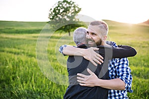 An adult hipster son with senior father on a walk in nature at sunset, hugging.