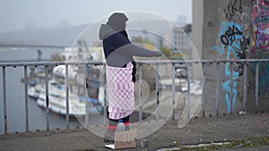 Adult homeless woman stay on the bridge near the river port in cold windy grey weather asking for alms