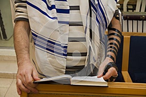 An adult jewish man praying with a tefillin on his arm, holding a bible book, while reading a pray