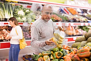 Adult latin american man choosing sweet pepper in supermarket with other buyer