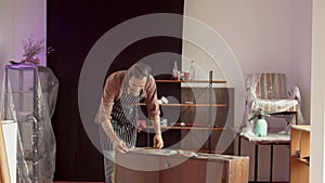 An adult male Caucasian furniture repairman in a special protective apron in a working workshop measures an old chest of