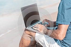 Adult male of mediterranean race using laptop while sitting on sandy beach. Freedom and travel concept